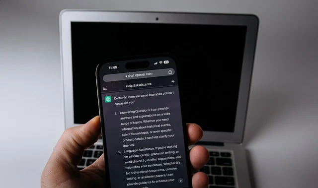 Una mano regge uno smartphone con una conversazione ChatGPT davanti a un computer portatile